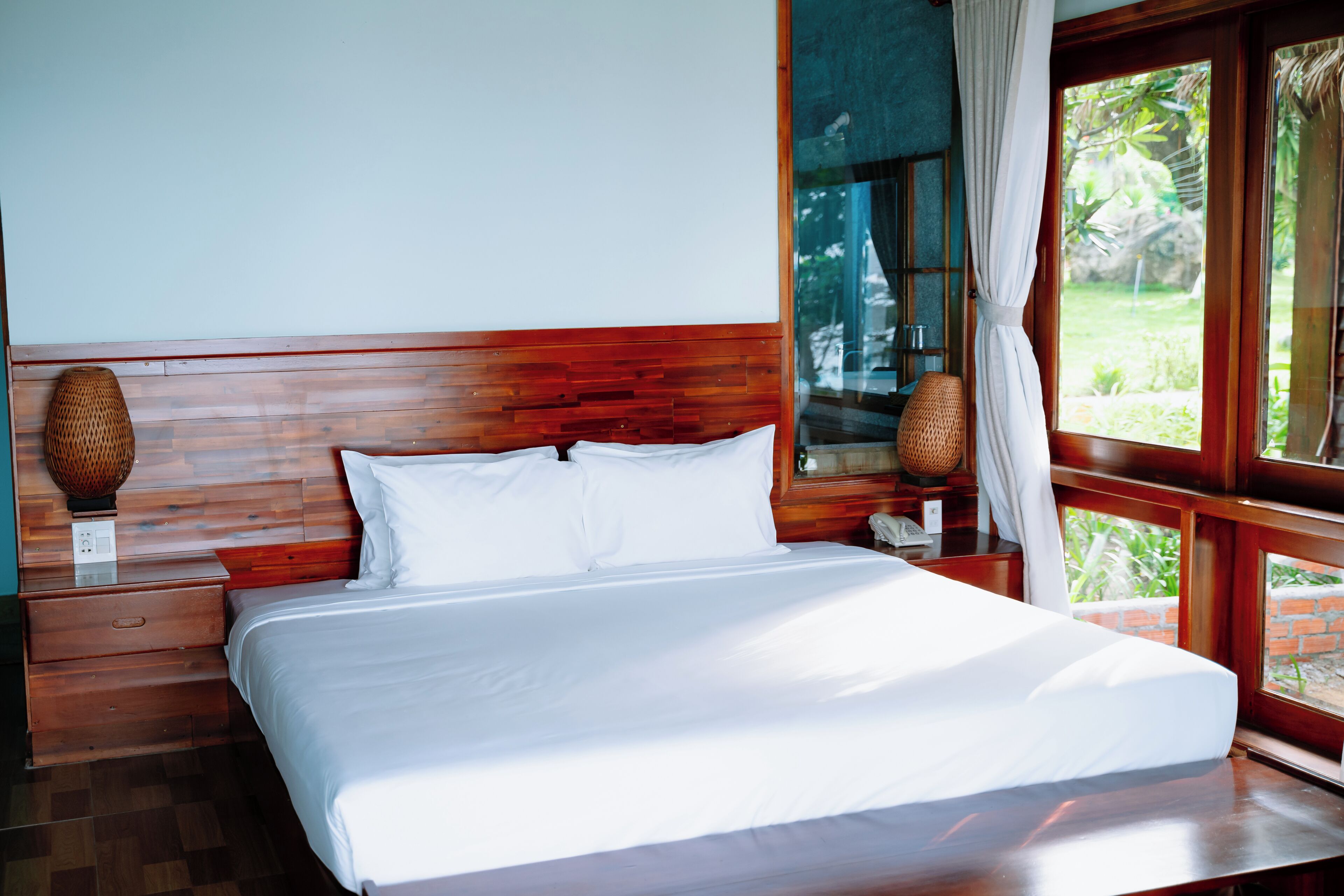 A neatly made bed with white linens in a room with wooden headboard and nightstands, large windows with curtains, and a view of greenery outside.