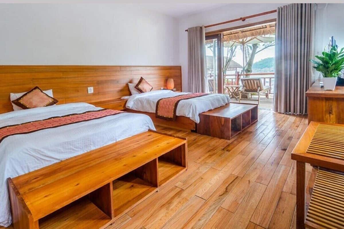 A hotel room with two wooden beds, a wooden bench, a desk, and a large window with a view of the outdoors.