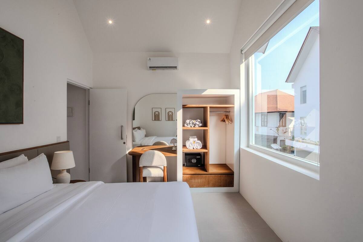 A modern hotel room with a large bed, a desk with a chair, a wardrobe, and a window with a view of buildings.
