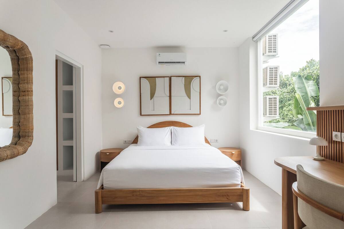A modern bedroom with a wooden bed, a large mirror, wall-mounted air conditioning, and a view of greenery outside.
