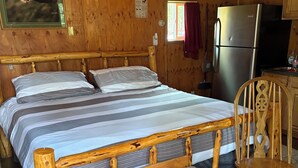 A wooden cabin bedroom with a large bed, a bench, a chair, and a kitchen area with a refrigerator.
