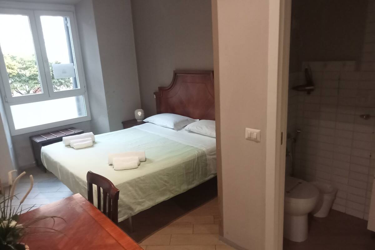 A hotel room with a bed, a wooden nightstand, a desk with a chair, a window, and a bathroom with a toilet and shower.