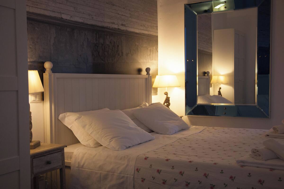 A neatly made bed with white linens and floral-patterned bedding, a wooden nightstand with a lamp, and a large mirror on the wall.