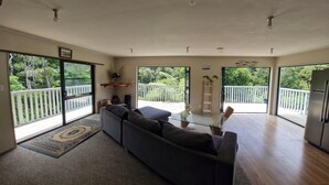 En rummelig opholdsstue med en stor, mørk sofa, et glasbord og udsigt til et hvidt hegn og grøntområde gennem store skydedøre.