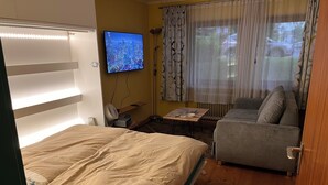 Ein Schlafzimmer mit einem Bett, einem Sofa, einem Fernseher, einem Tisch mit Ventilator und einem Fenster mit Vorhängen.