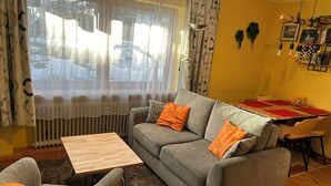 Ein Wohnzimmer mit grauem Sofa, orangefarbenen Kissen, einem Holztisch und einem Essbereich mit rotem Tischlein.
