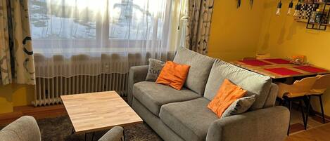 A living room with a grey sofa, orange cushions, a wooden coffee table, and a dining area with a red tablecloth.