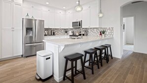 Een moderne keuken met witte kasten, een roestvrijstalen koelkast, een ingebouwde magnetron, een groot kookeiland met barstoelen en hanglampen.