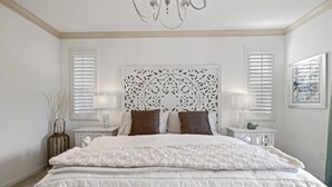 Een slaapkamer met een groot bed, een decoratief hoofdeinde, twee nachtkastjes met lampen en een raam met jaloezieën.
