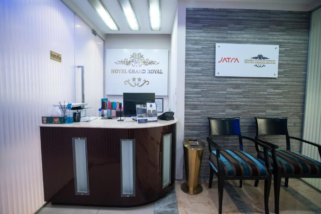 Un comptoir d’accueil avec une enseigne indiquant « Hôtel Grand Royal » et « JATCA Hôtel Grand Royal ». Devant le comptoir, on trouve deux chaises dotées de coussins à rayures.