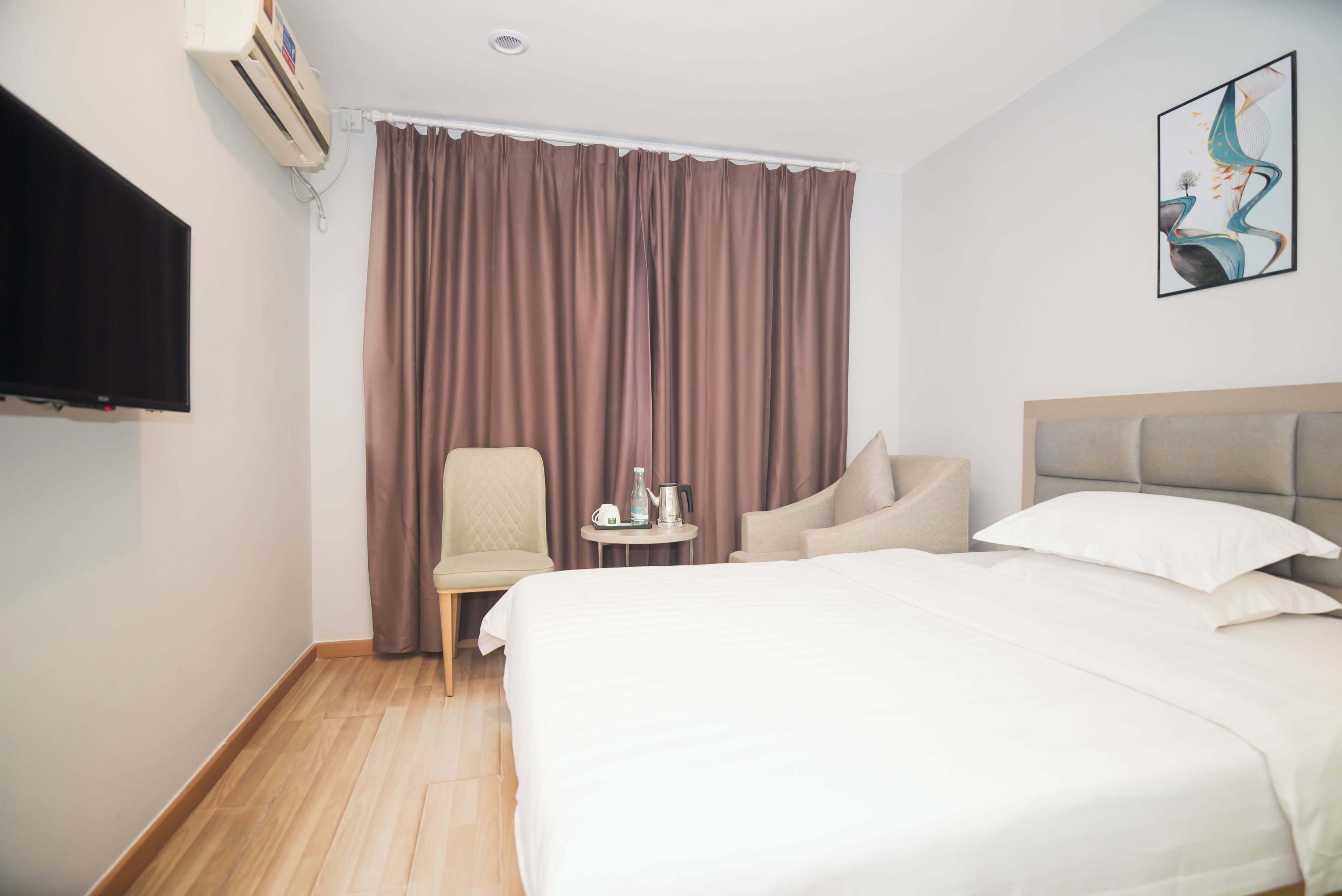 A hotel room with a large bed, a flat-screen TV, a chair, a small table with a kettle, and a painting on the wall.