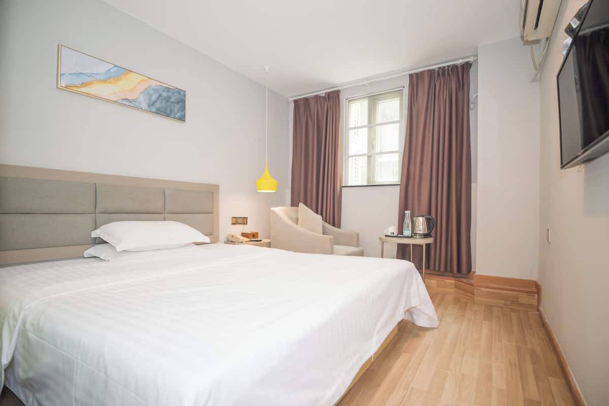 A hotel room with a large bed, a chair, a small table with a lamp, a painting on the wall, and a window with curtains.