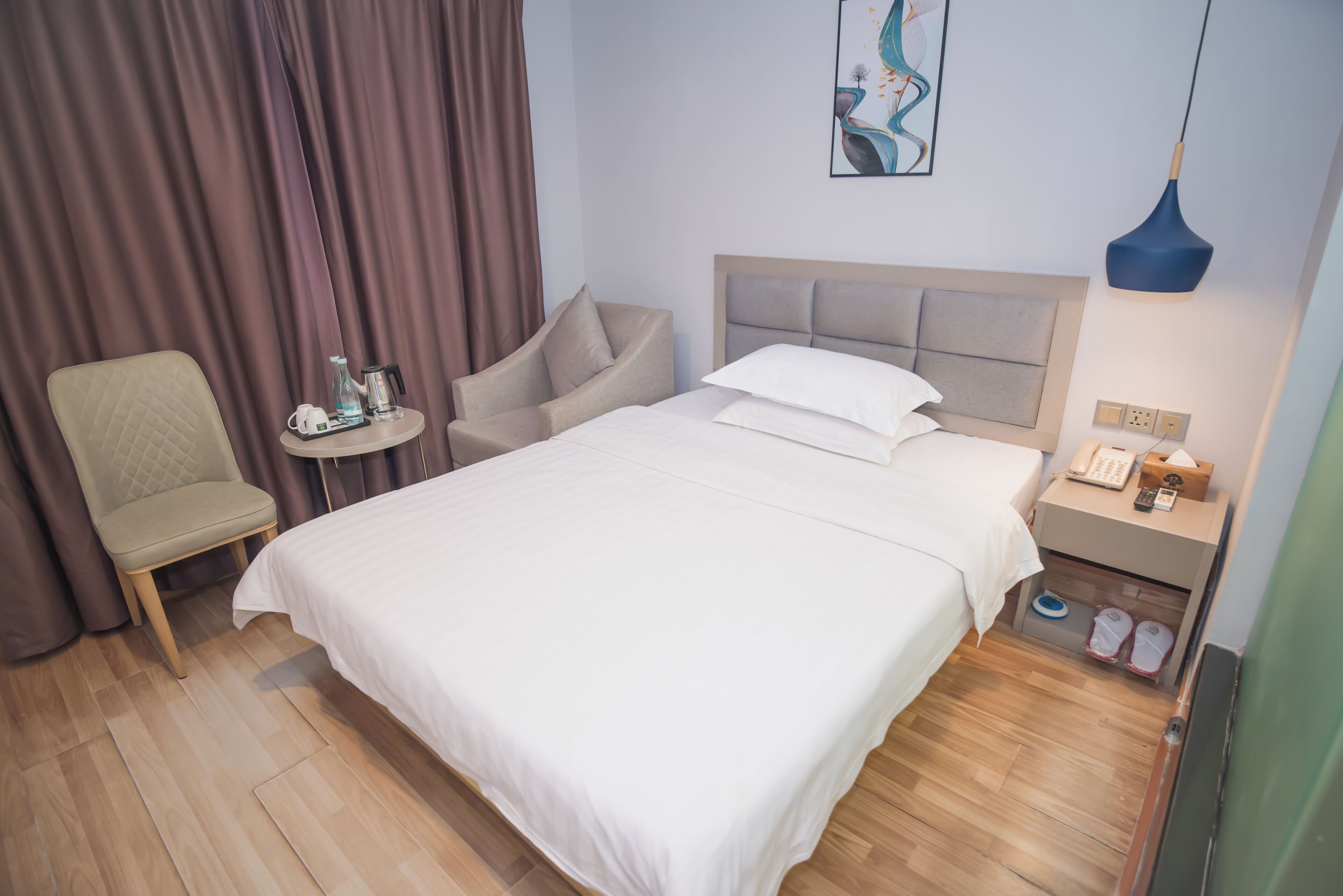 A hotel room with a large bed, a chair, a small table with a phone and a bottle, a nightstand with a lamp, and a painting on the wall.