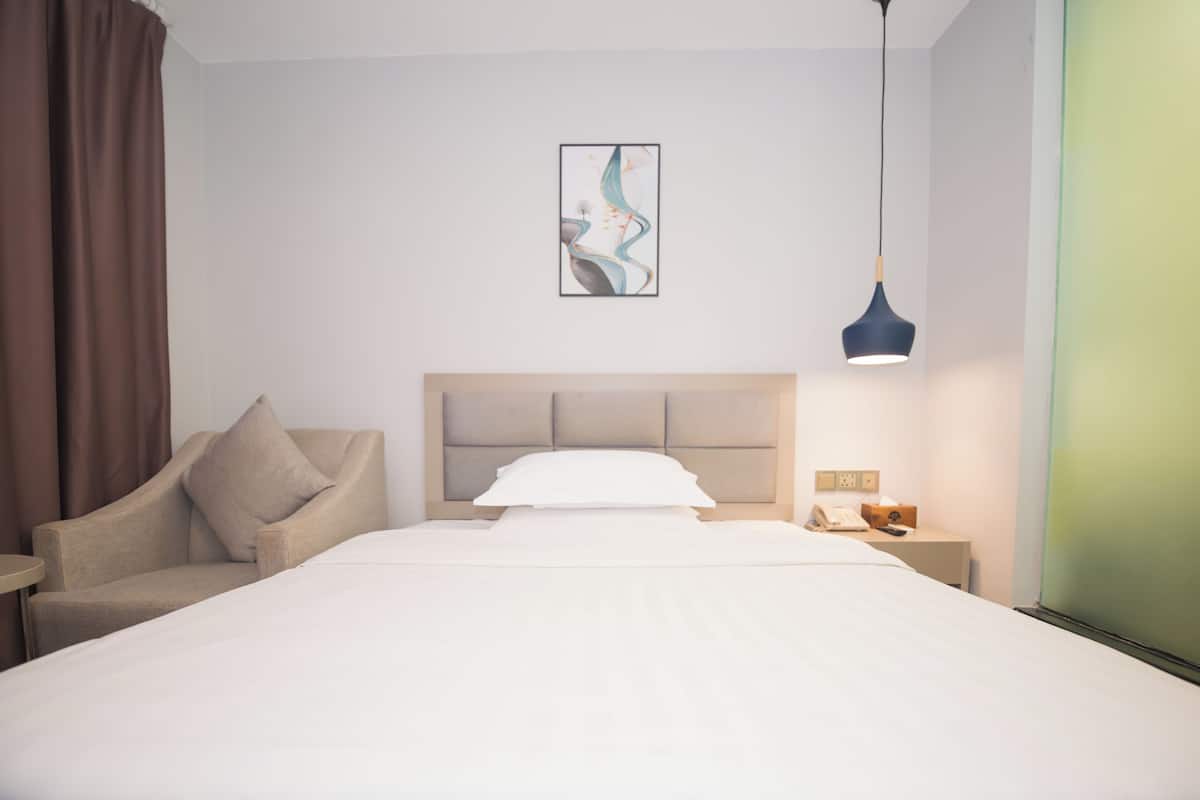 A hotel room with a bed, a chair, a bedside table with a lamp, and a painting on the wall.