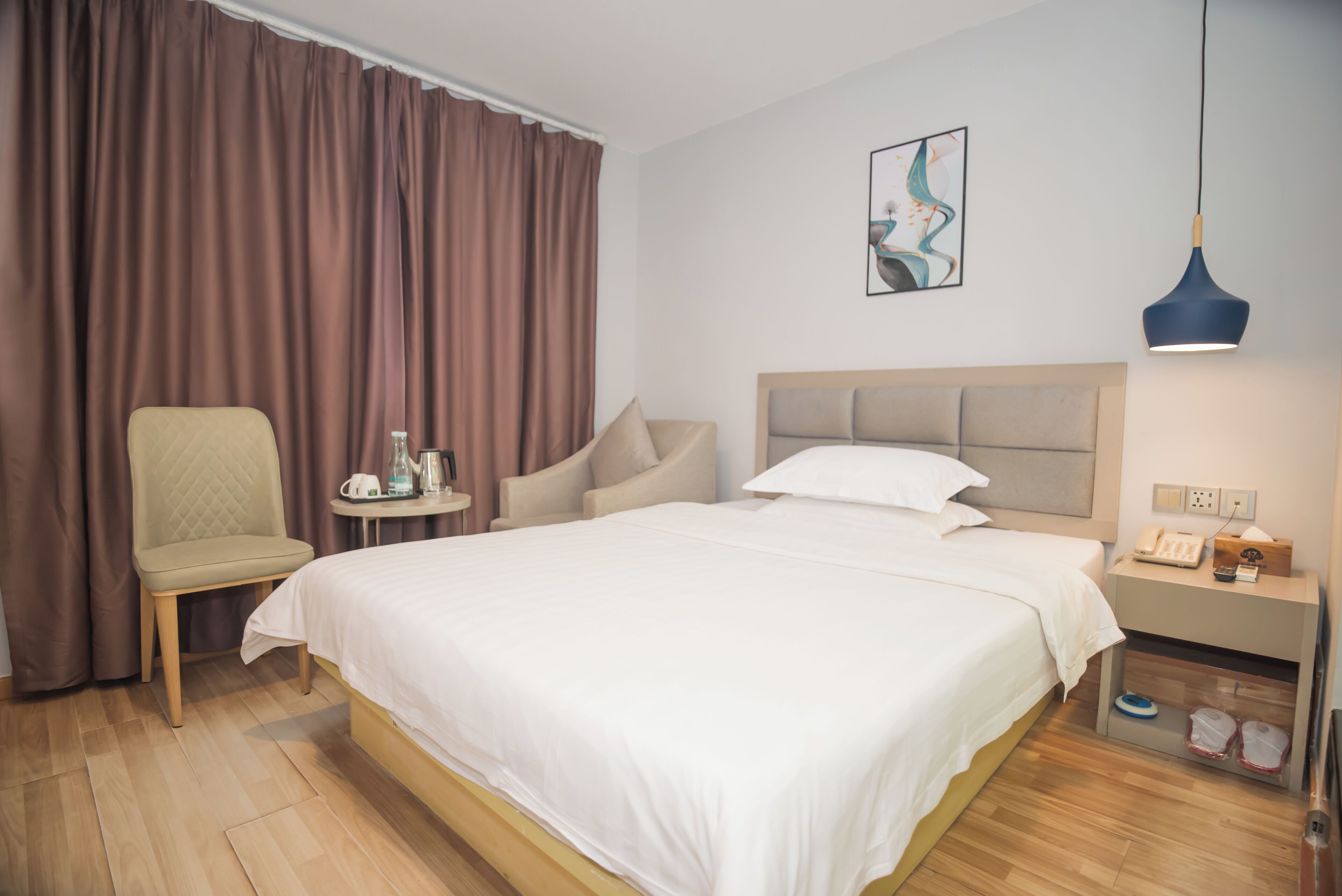 A hotel room with a large bed, a chair, a small table with a kettle, a nightstand with a lamp, and a painting on the wall.