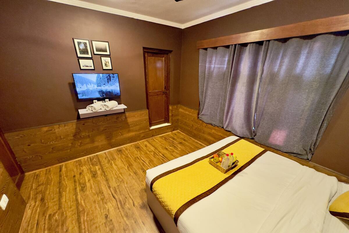 A hotel room with a wooden floor, a bed with a yellow and white bedspread, a television on the wall, and a door with a wooden frame.