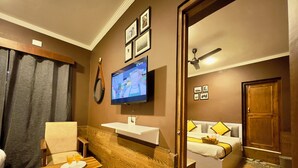 A hotel room with a wooden floor, a TV mounted on the wall, a bed with a white and yellow bedspread, a wooden bench, and a small table with a glass of orange juice. - Hotel Jafson Accommodates (Srinagar)