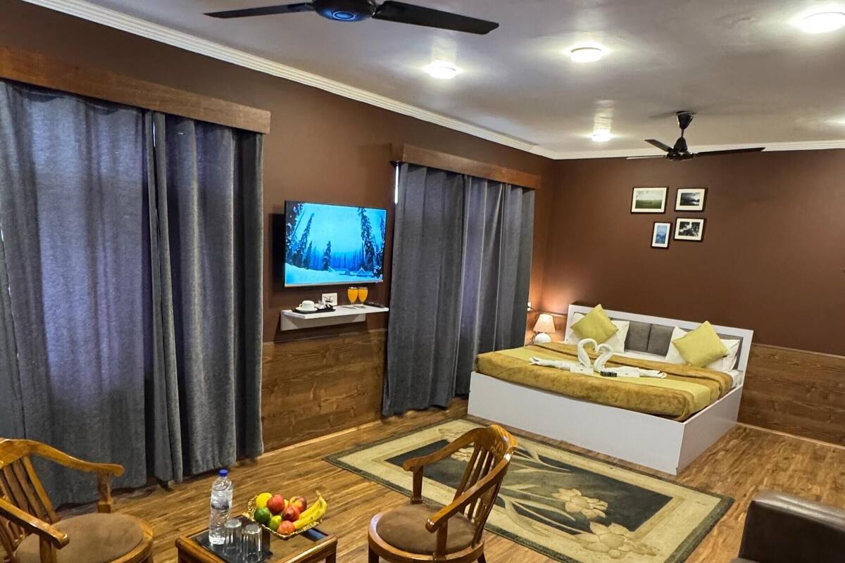 A modern living room with a sofa, two wooden chairs, a coffee table with fruit and glasses, a TV, and a ceiling fan.