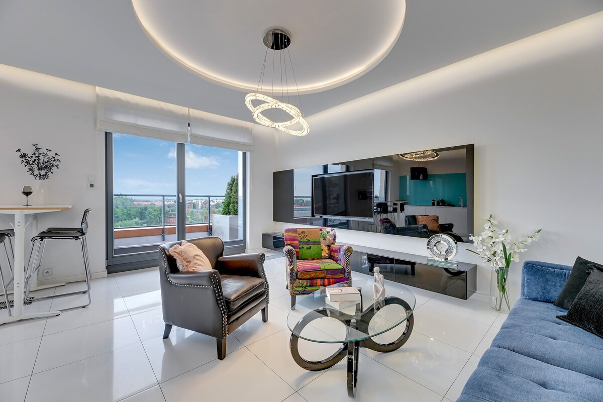 Uma sala de estar moderna com uma televisão grande, um sofá, uma poltrona colorida e uma mesa de centro de vidro.