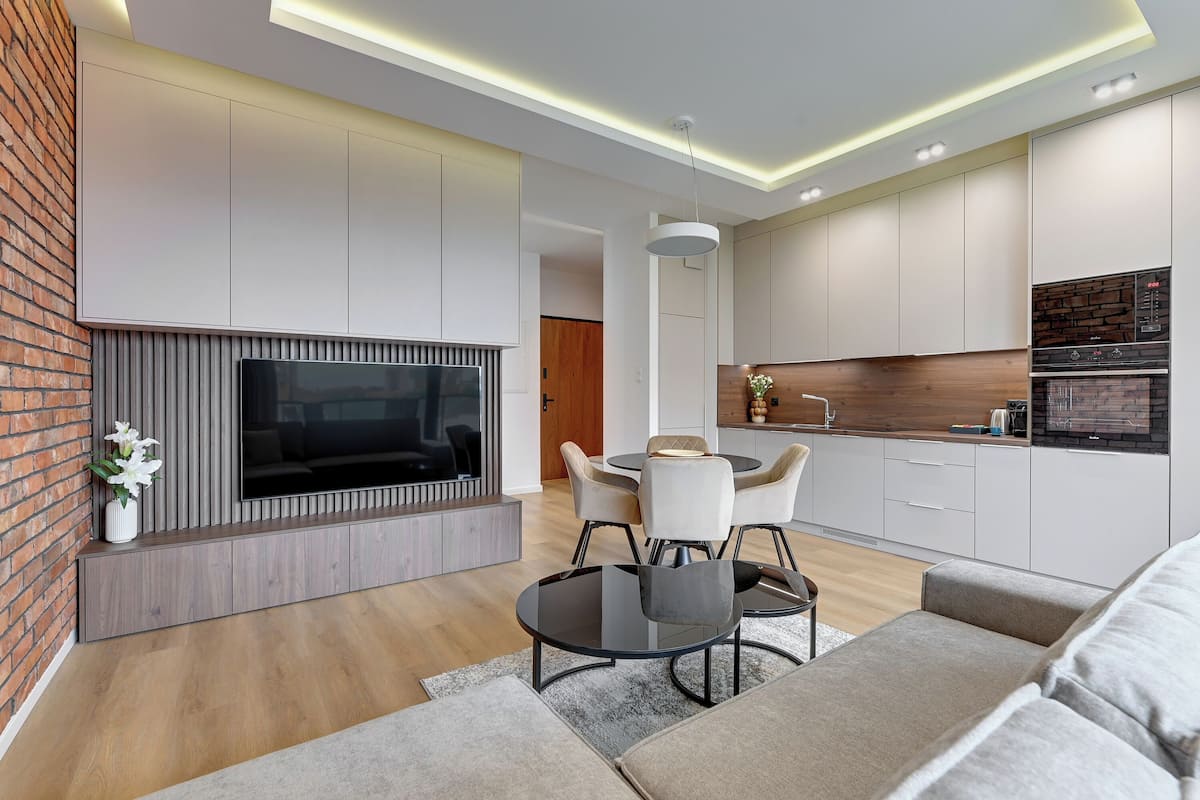 Una sala de estar moderna con un televisor de pantalla plana, un comedor con una mesa redonda y sillas, y una cocina con gabinetes blancos y un salpicadero de ladrillo.