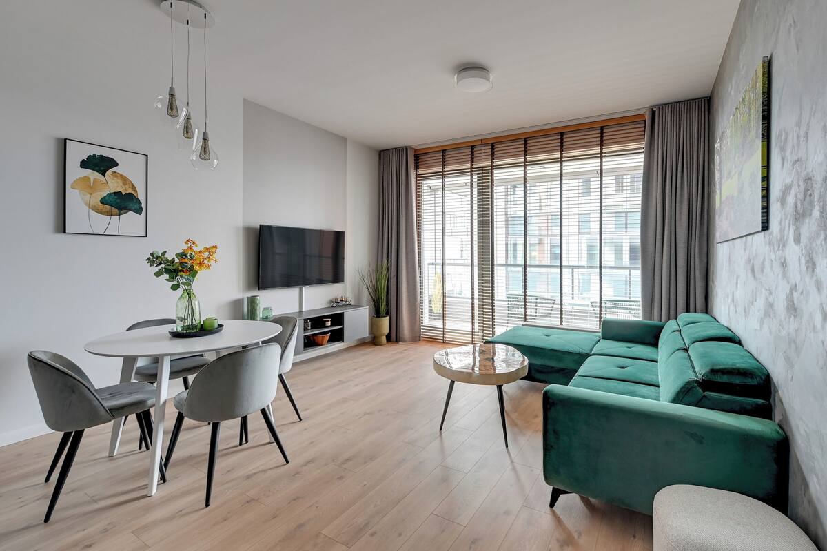 Una sala de estar moderna con un sofá verde, una mesa de centro redonda, una mesa de comedor con sillas, un televisor de pantalla plana y un amplio ventanal con persianas.