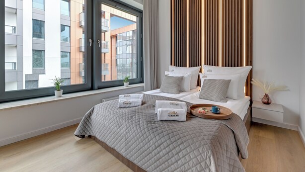 Moderni hotellihuone, jossa on suuri sänky, yöpöytä, yövalaisin, ikkuna, josta on näkymä kaupunkiin, sekä koristeellinen seinäpaneeli.