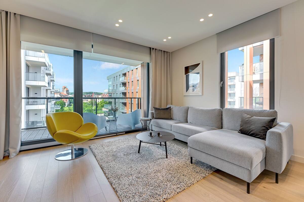 Una sala moderna con un sofá gris, un sillón amarillo, una mesa de centro y amplias ventanas con vistas a edificios y zonas verdes.
