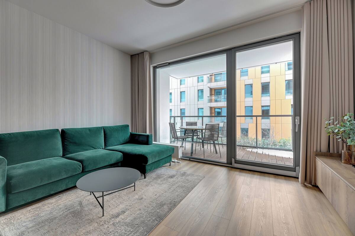 Una sala moderna con un sofá verde, una mesita de centro y un ventanal corredero que da a un balcón exterior con vistas a un edificio.