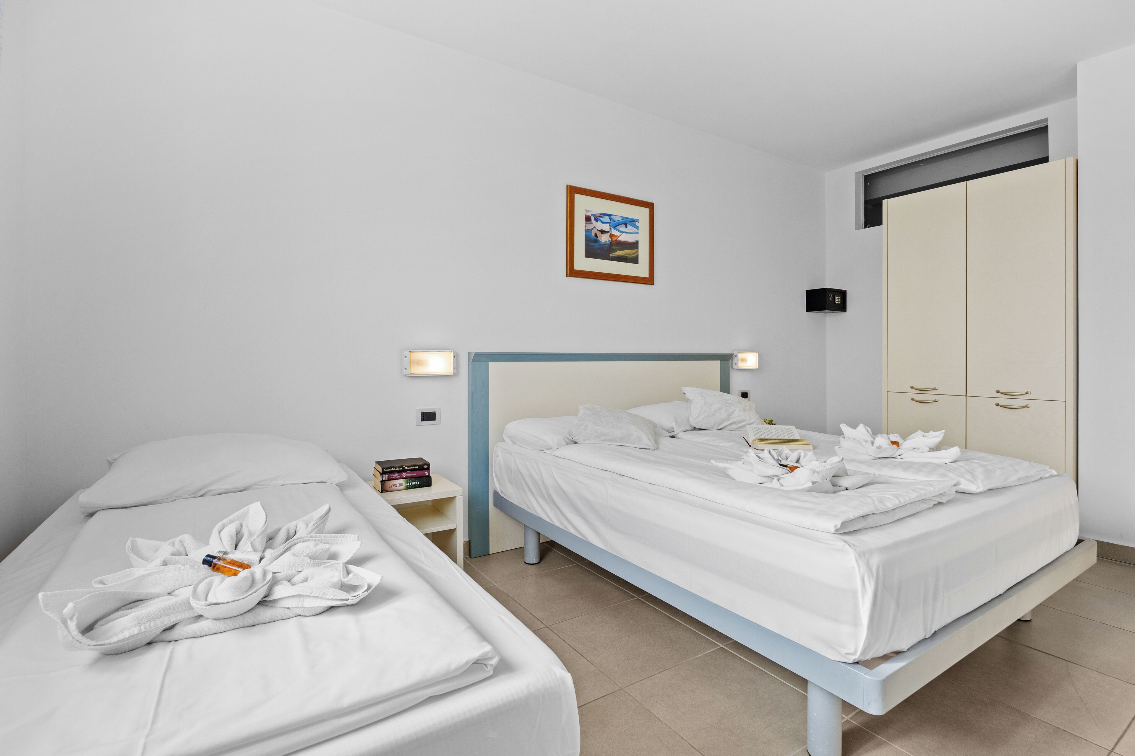 Habitació d'hotel amb dos llits, un armari, una taula de nit amb una làmpada i un quadre al mur.