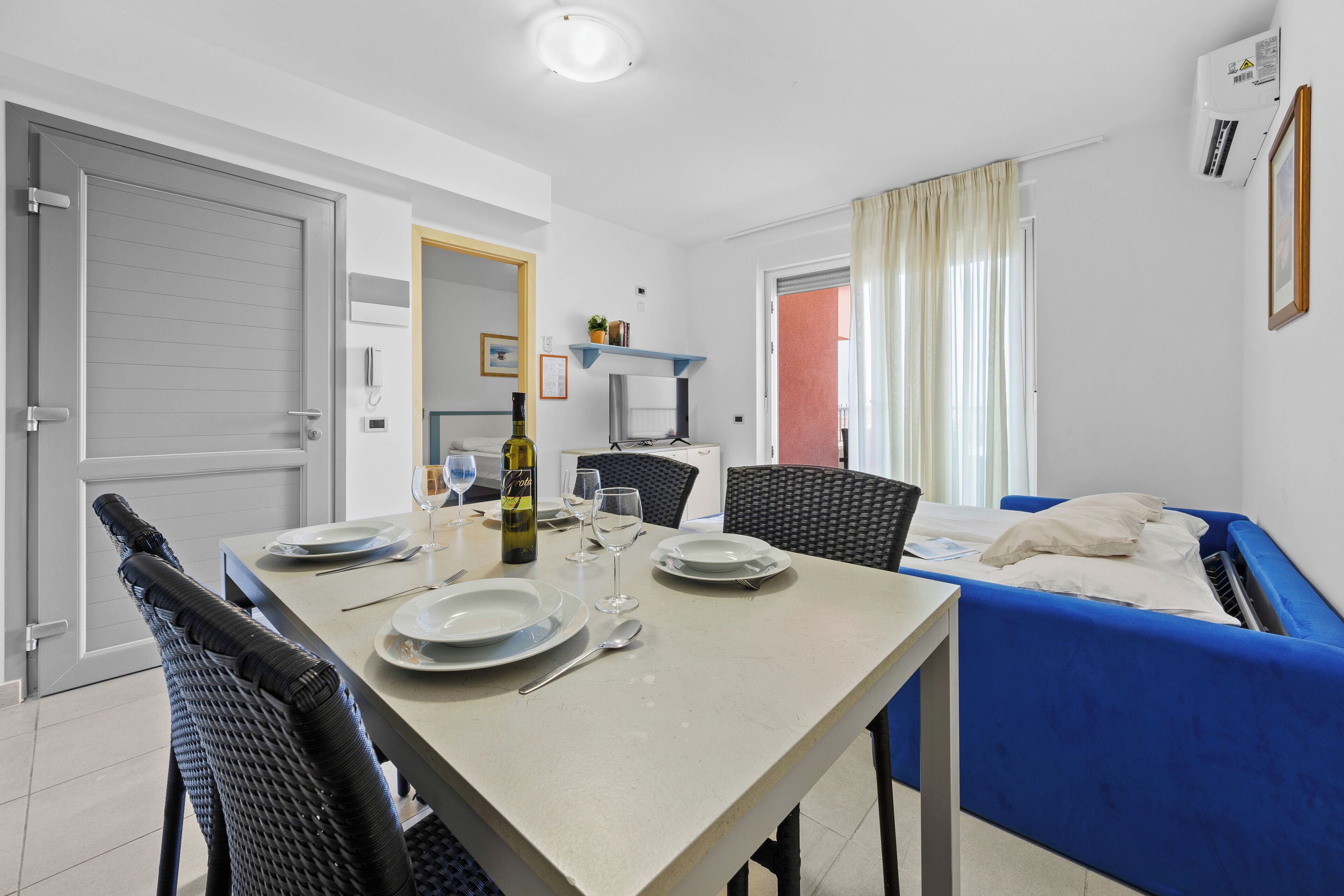 A room with a dining table set for two, a blue sofa, a bed, and a small shelf with a plant.