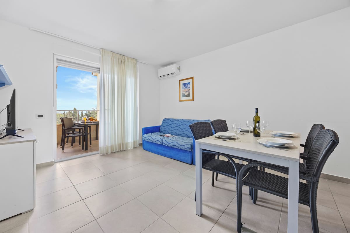 A modern living room with a dining area, a blue sofa, a white table, and a balcony with a view.