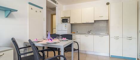 Une cuisine compacte avec des meubles blancs, un four à micro-ondes et un petit réfrigérateur. On y trouve une table à manger avec des chaises, ainsi qu’une chaise bleue.