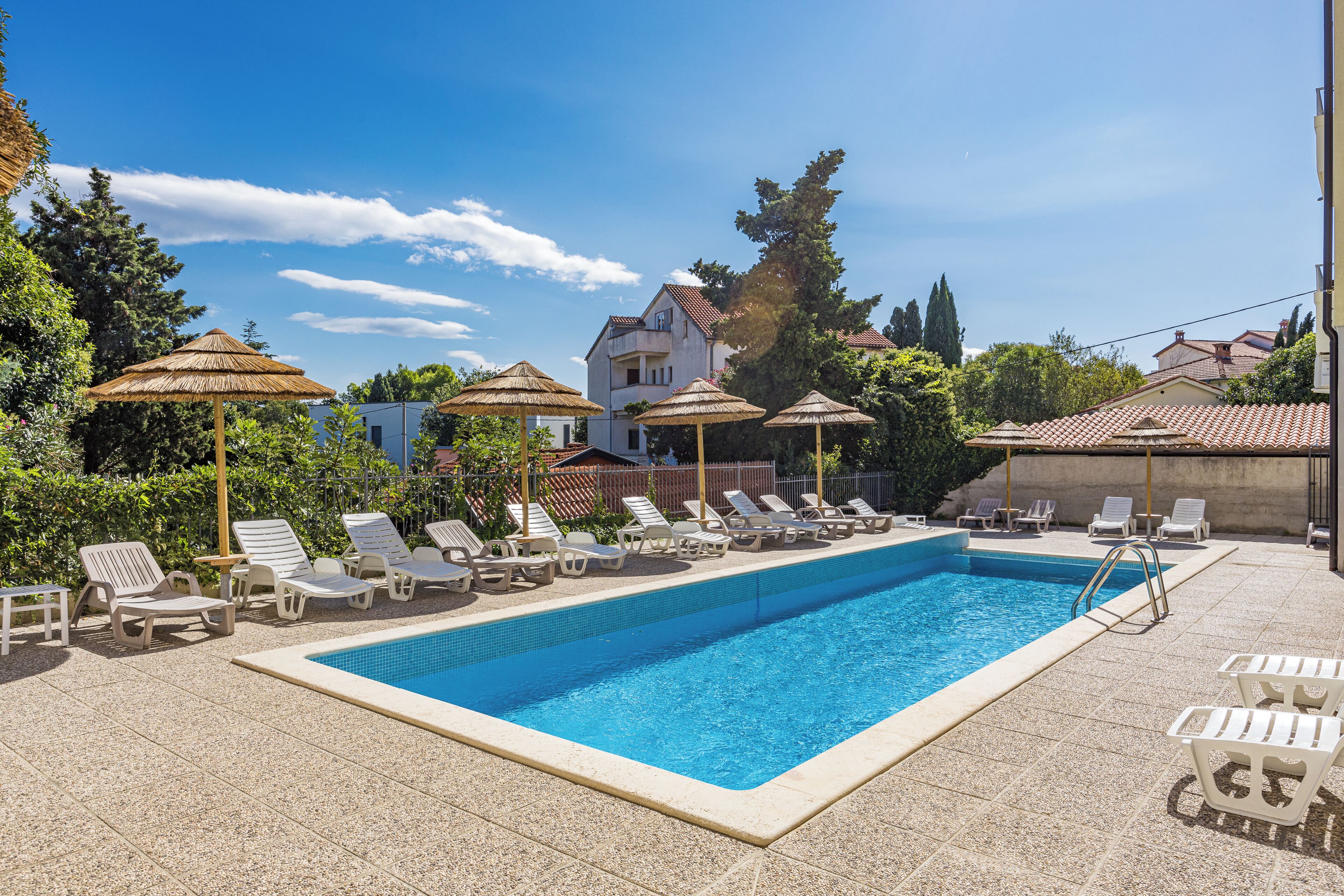Una piscina rectangular con borde de azulejos, rodeada de sillas de descanso y sombrillas de paja.
