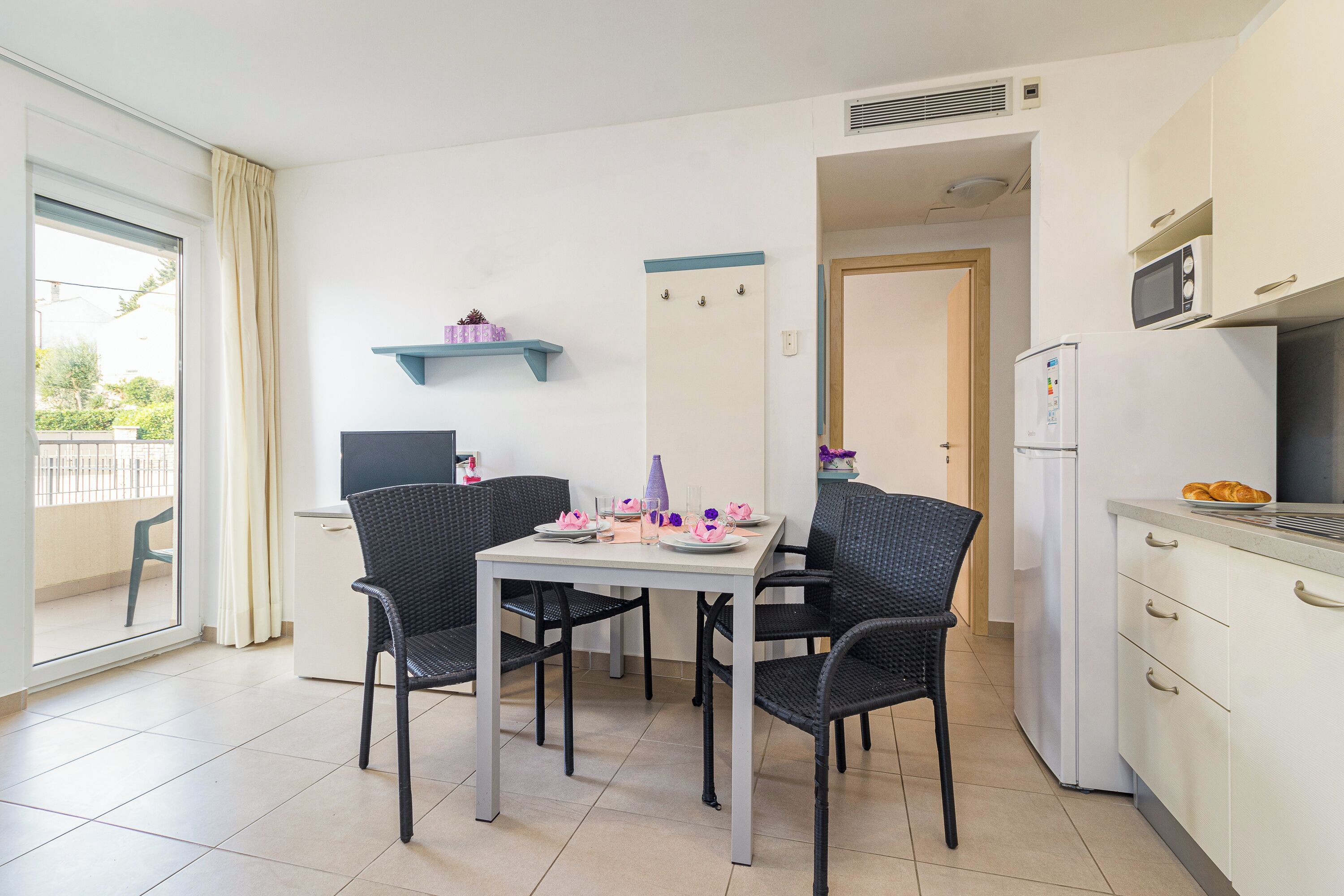 A small, modern apartment with a dining area featuring a white table and black chairs, a kitchen with white cabinets and a microwave, and a sliding glass door leading to a balcony.