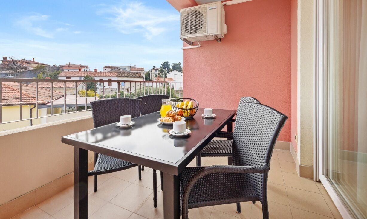 Un balcón con mesa lista para el desayuno, sillas de mimbre y vistas a edificios distantes.