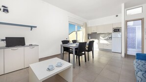 Une pièce moderne et bien éclairée, comprenant une table à manger, des chaises, un téléviseur posé sur un meuble, un coin cuisine équipé d’appareils électroménagers et une vue par la fenêtre.