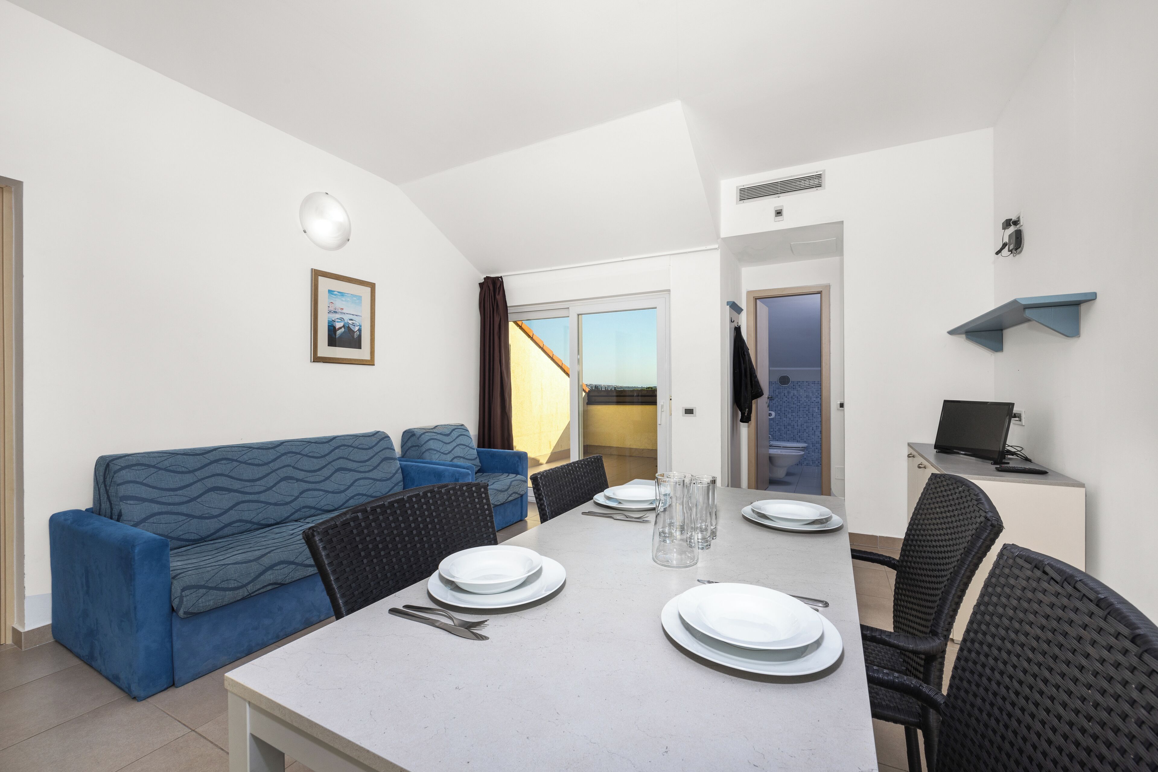 Un espace de repas moderne avec une table dressée pour quatre personnes, un canapé bleu et un ordinateur portable posé sur un petit bureau.