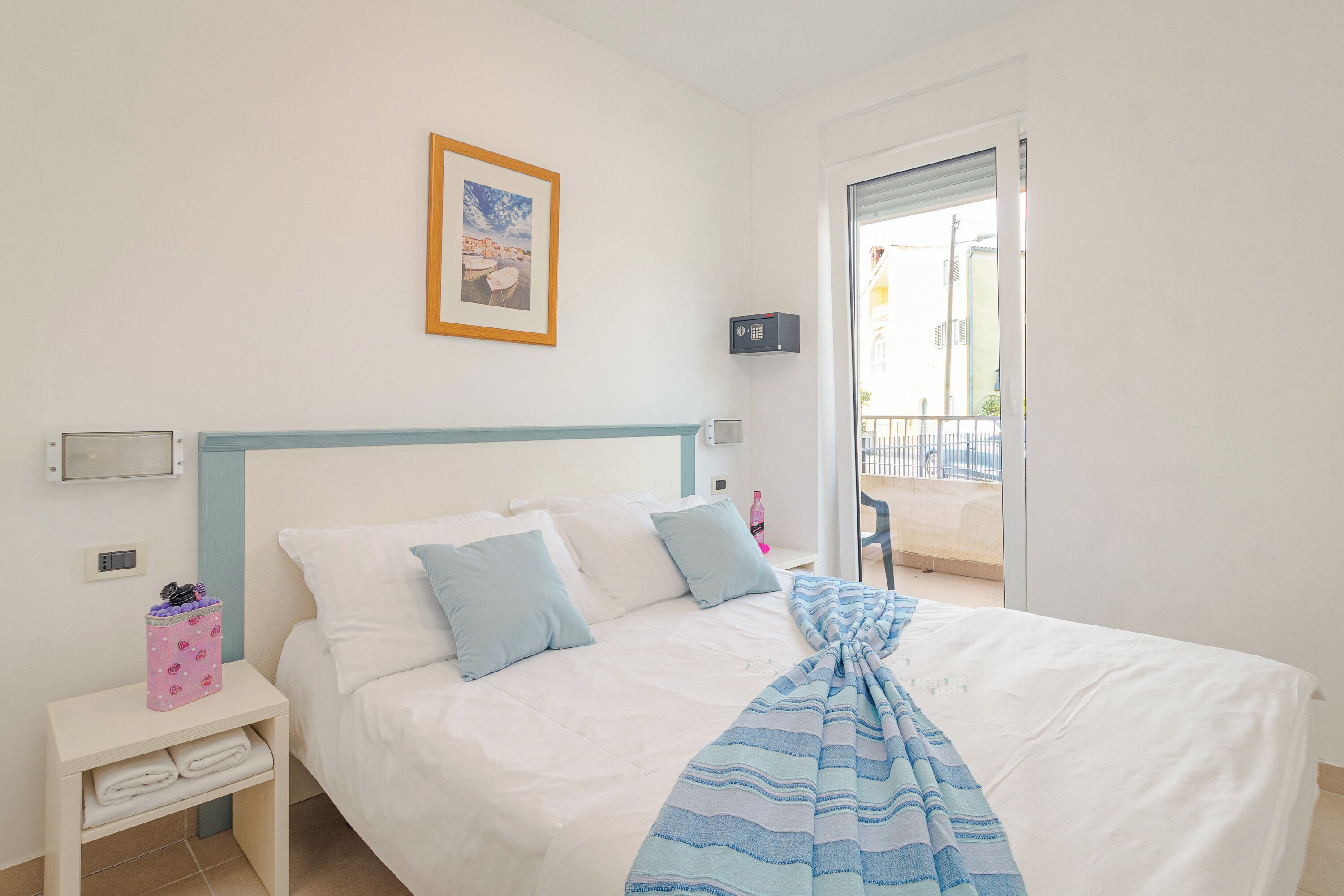 A neatly made bed with a striped blanket, a bedside table with a pink basket, and a framed picture on the wall.