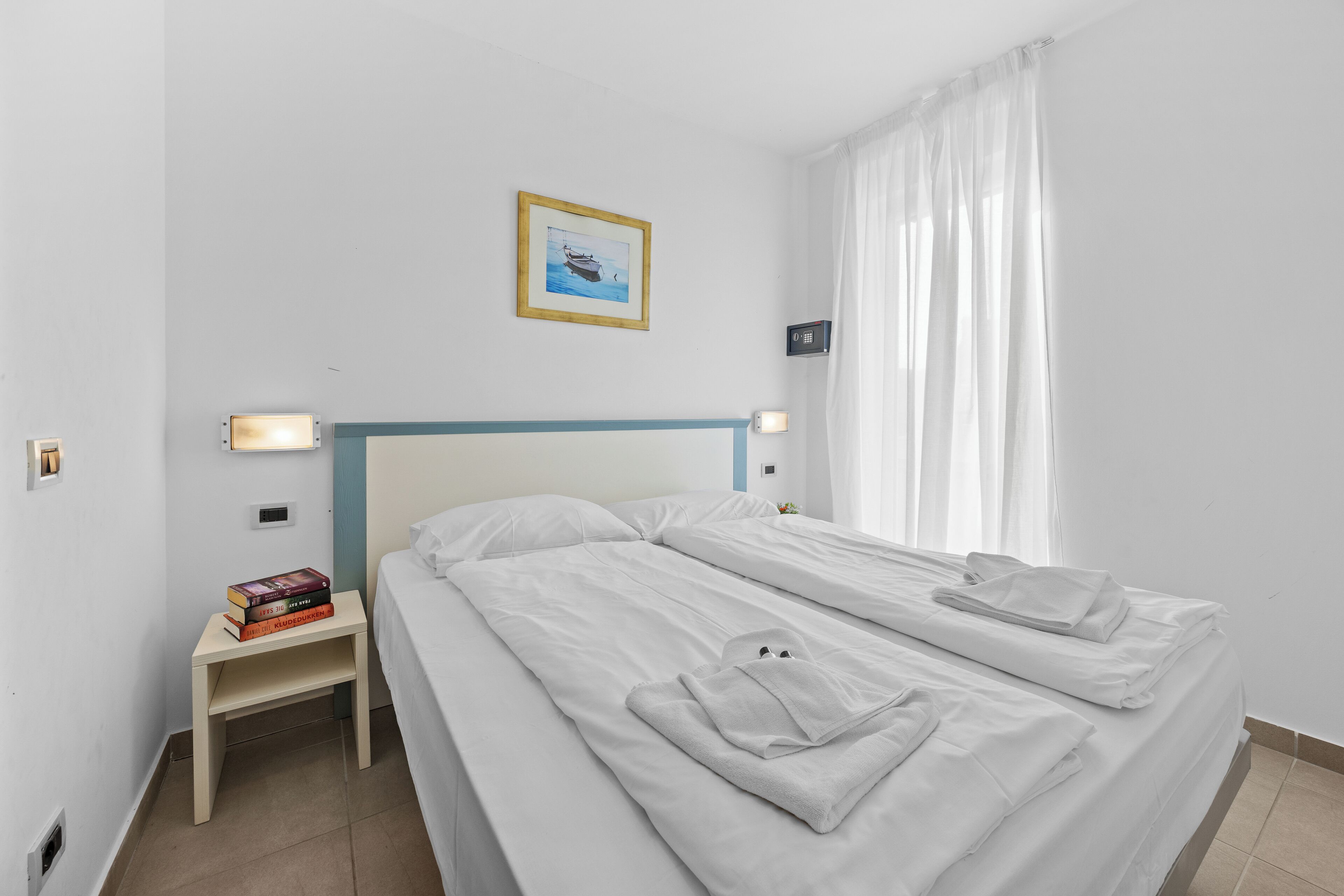 A hotel room with two beds, a nightstand with books, a framed picture on the wall, and a window with curtains.
