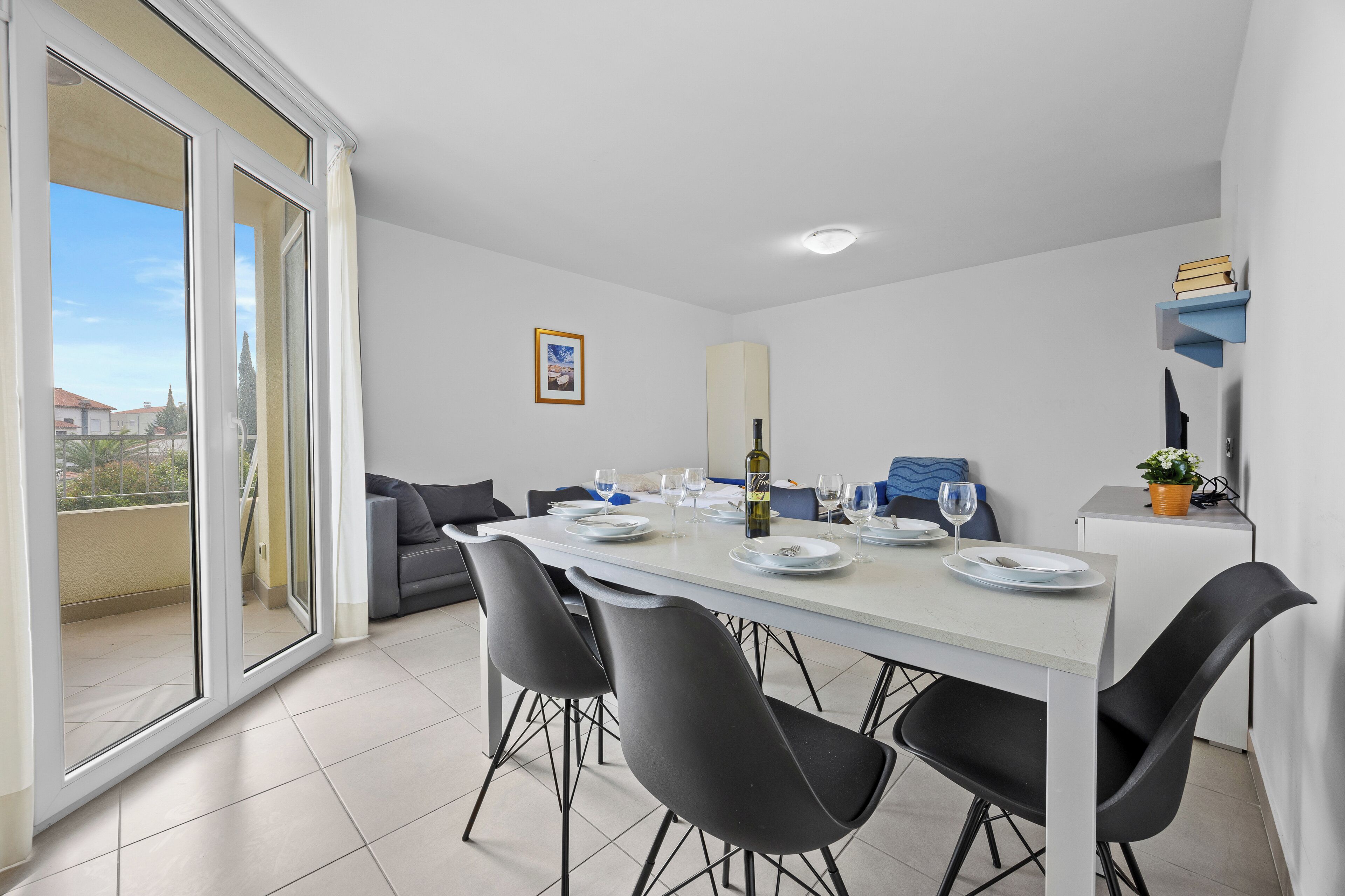 A dining area with a table set for four, black chairs, a white table, and a view through a sliding glass door to the outside.
