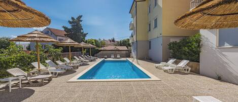 A swimming pool with sun loungers and thatched umbrellas, surrounded by buildings.