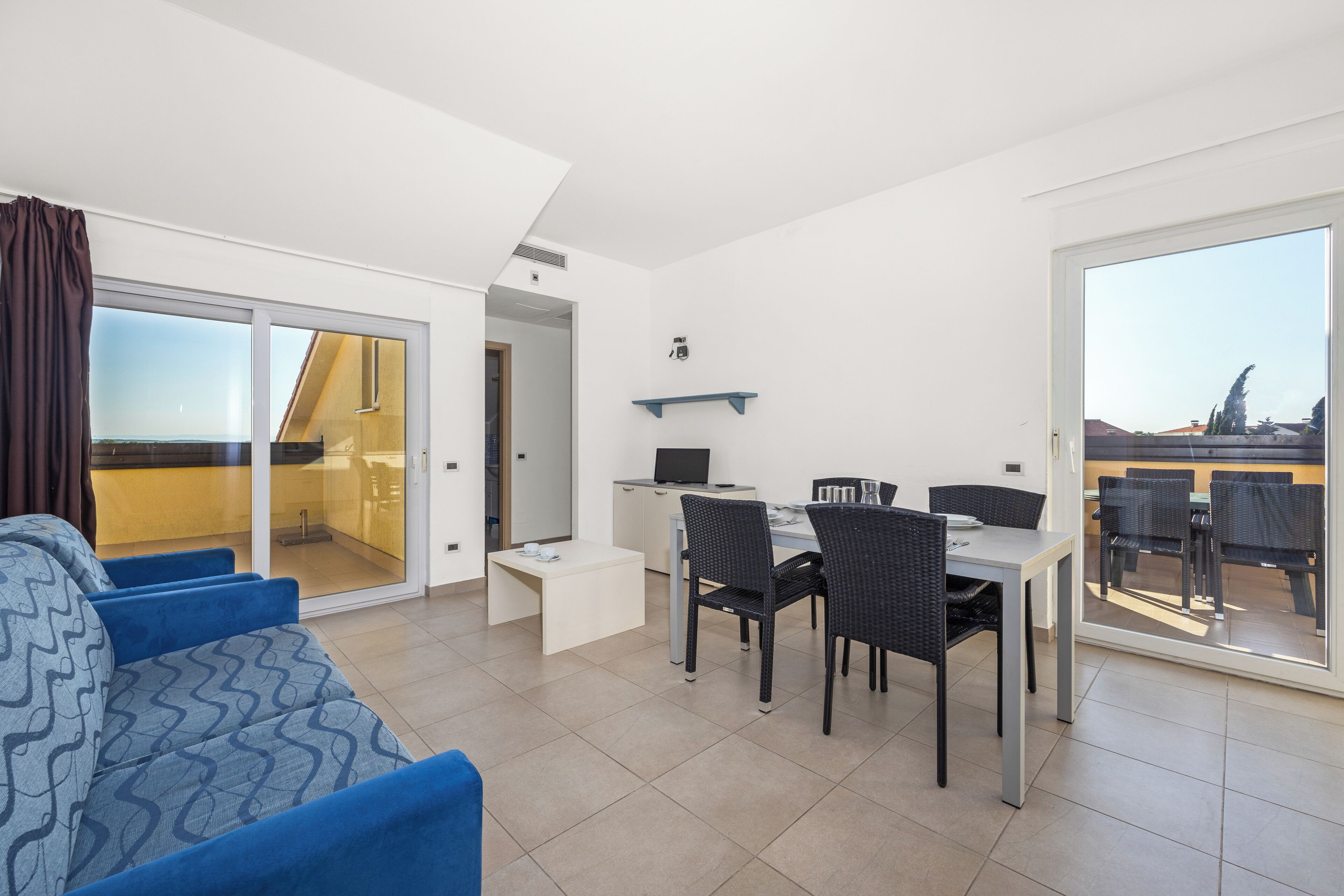 Un salon moderne avec un canapé, une table à manger et des chaises. Une porte-fenêtre coulissante donne accès à un balcon avec vue.
