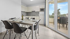 Une cuisine moderne avec des meubles blancs, une table à manger pour quatre personnes et une vue sur l’extérieur grâce à une porte-fenêtre coulissante.
