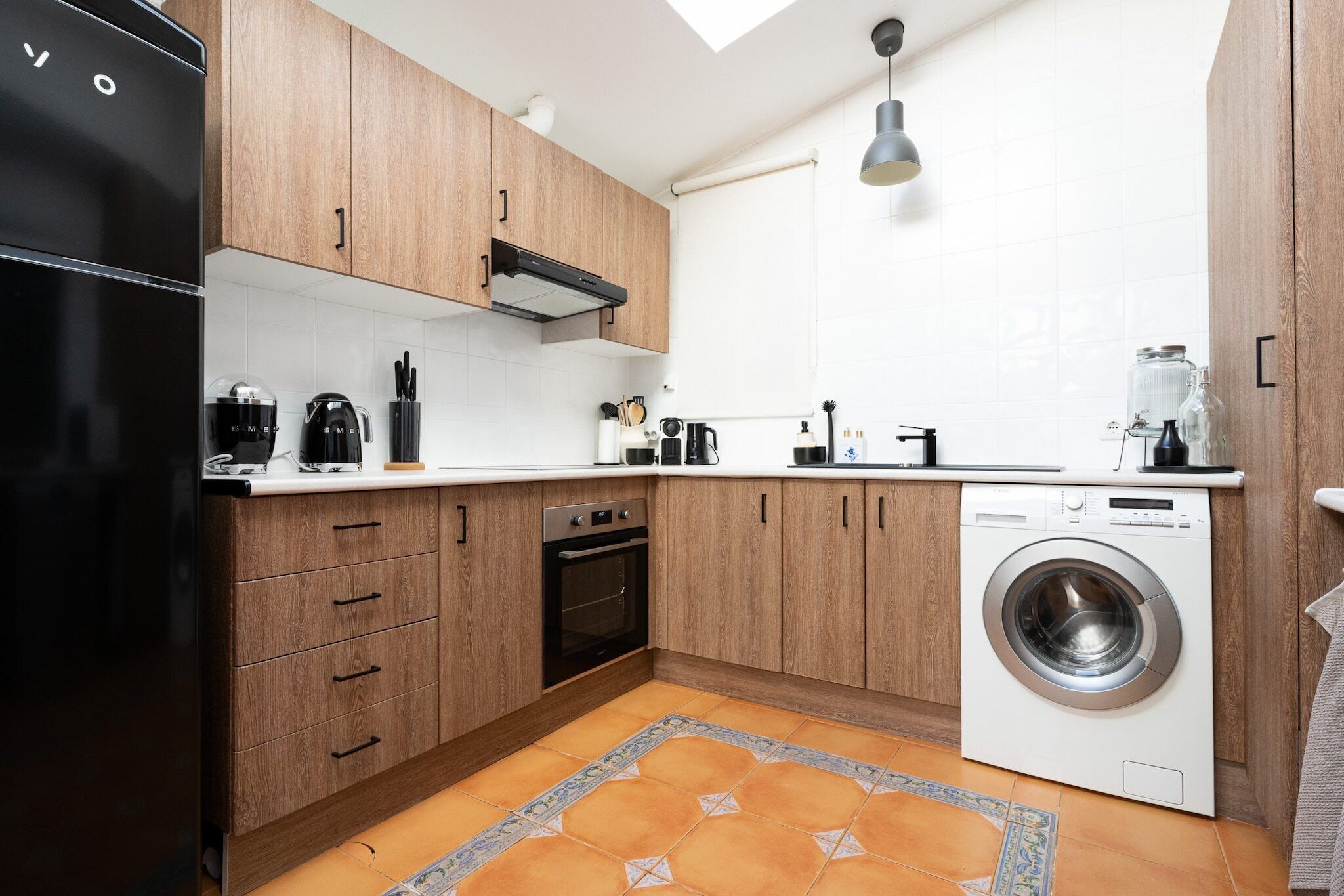 A compact kitchen with wooden cabinets, a black refrigerator, a built-in oven, a washing machine, and a tiled backsplash.