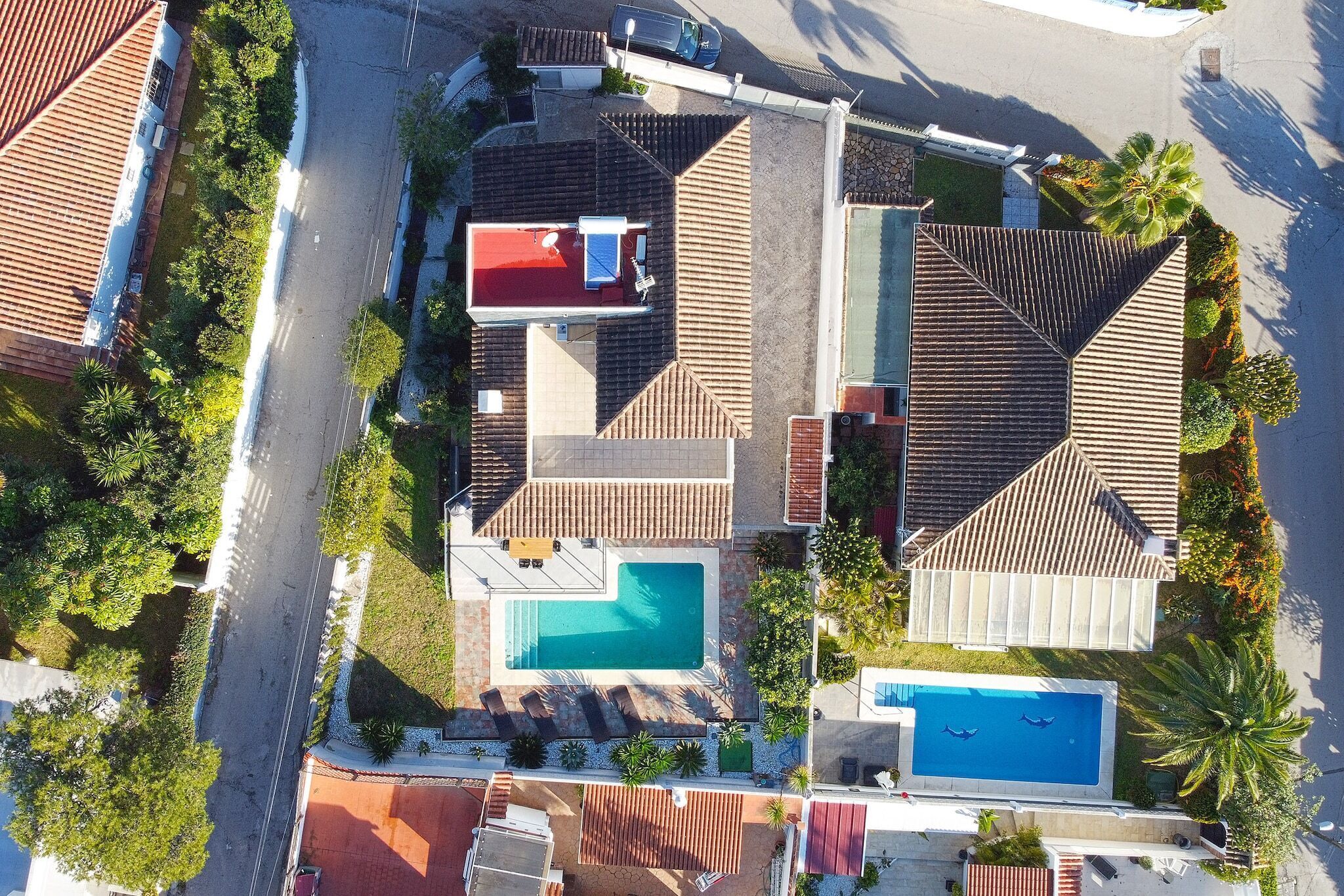 An aerial view of a residential area with two swimming pools, a red-roofed building, and a large house with a sloped roof.