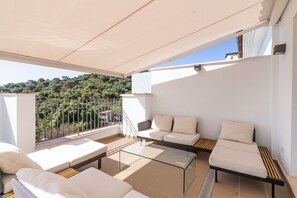 A balcony with a glass table, white sofas, and a view of a green hillside.