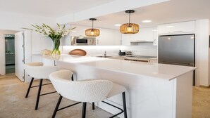 A modern kitchen with white cabinetry, a large island with bar stools, a stainless steel refrigerator, and a microwave.