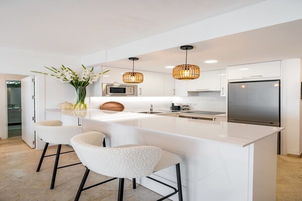 A modern kitchen with white cabinetry, a large island with bar stools, a stainless steel refrigerator, and a microwave. - Seaside Stay with Pool (Estepona)