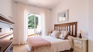 Ein Schlafzimmer mit Bett, Nachttisch, Kommode und Blick ins Freie.