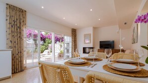 Un coin repas avec une table dressée pour quatre personnes, des chaises en osier, un téléviseur et une vue sur un jardin parsemé de fleurs roses.